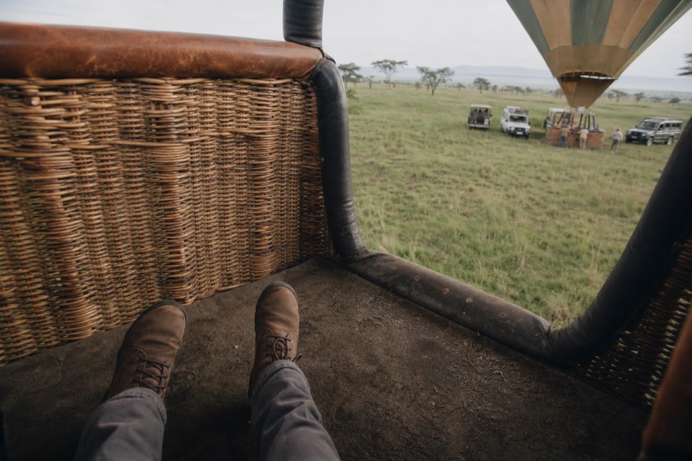 A passenger's dusty boots planted apart on the matted floor of a balloon basket compartment, low morning shadow falling across the weave
