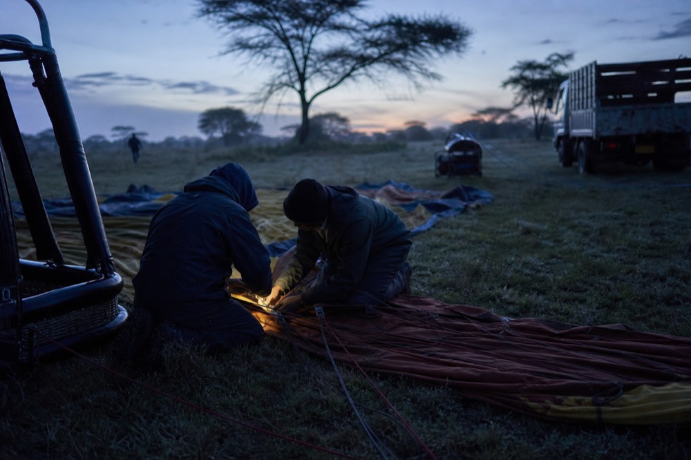 Ground crew crouched over rigging lines and a folded envelope laid out on wet grass in blue pre-dawn light, checking metal connections by torchlight