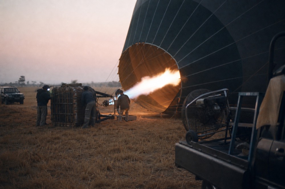Propane burner firing a long orange flame into a balloon mouth before dawn, crew in headlamps steadying the basket on dry grass