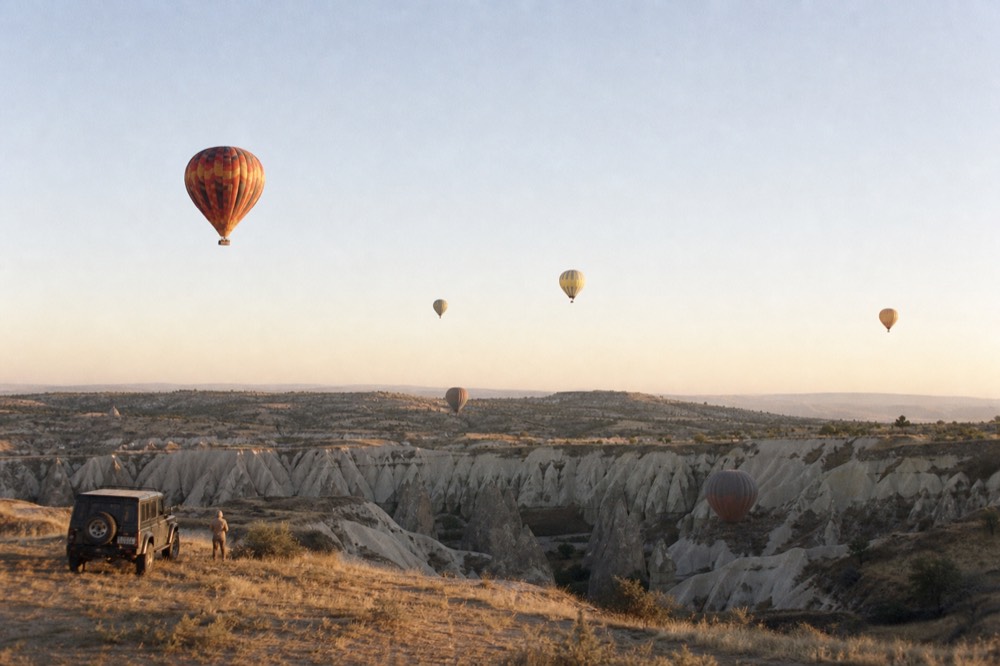 Several balloons at different heights above eroded rock valleys at sunrise, warm light striking the pale stone below them