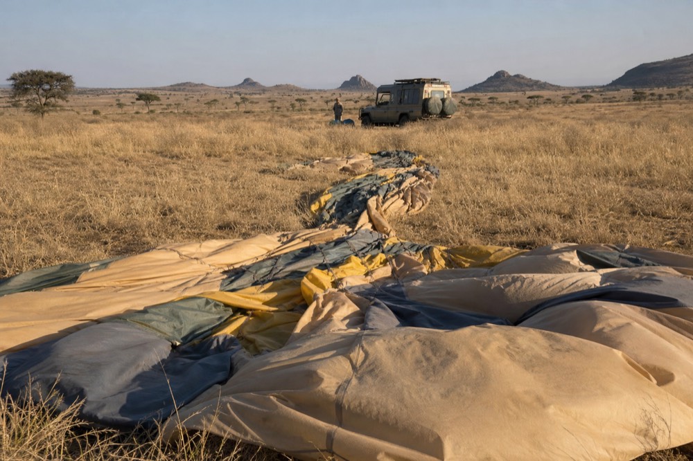 A deflated balloon envelope spread flat over trampled golden grass after landing, drag marks pressed through the stems, recovery truck waiting far behind