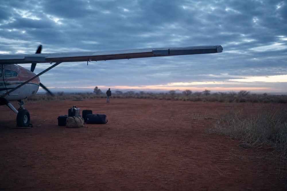 A small propeller aircraft parked on a red dirt airstrip fringed with dry grass under morning haze, luggage stacked beside the wing