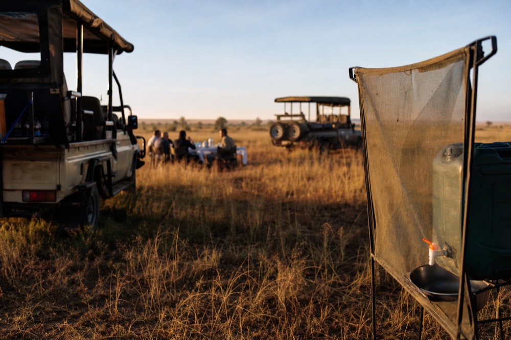 A canvas-screened washstand and a jerrycan tap standing in long yellow grass beside parked safari vehicles at a bush breakfast site in morning sun