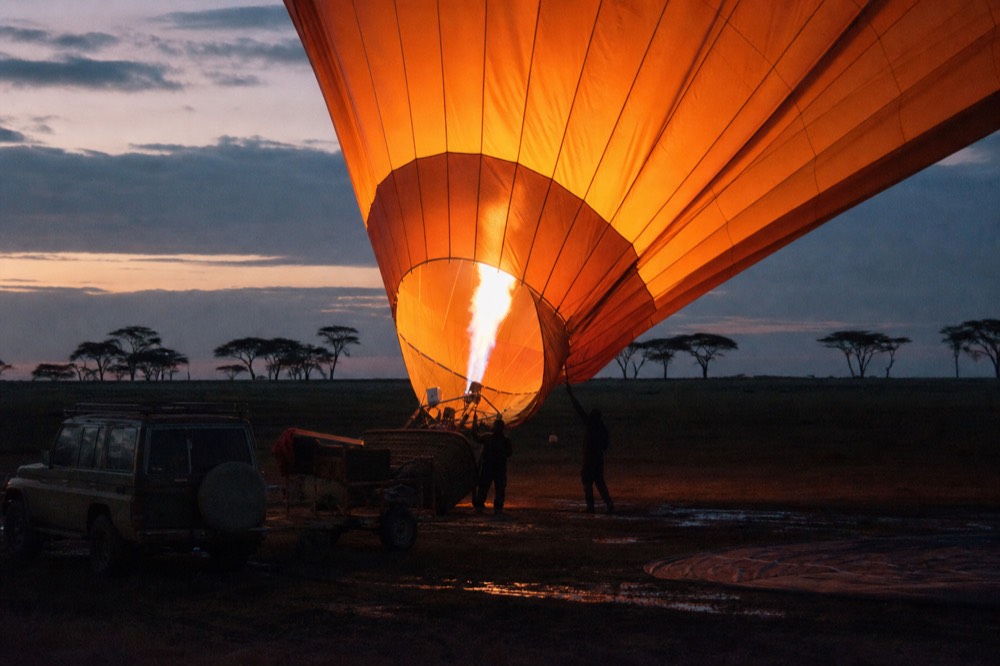 Burner flame lighting the inside of a balloon envelope from below before dawn, orange glow across the nylon panels, dark wet plains beyond