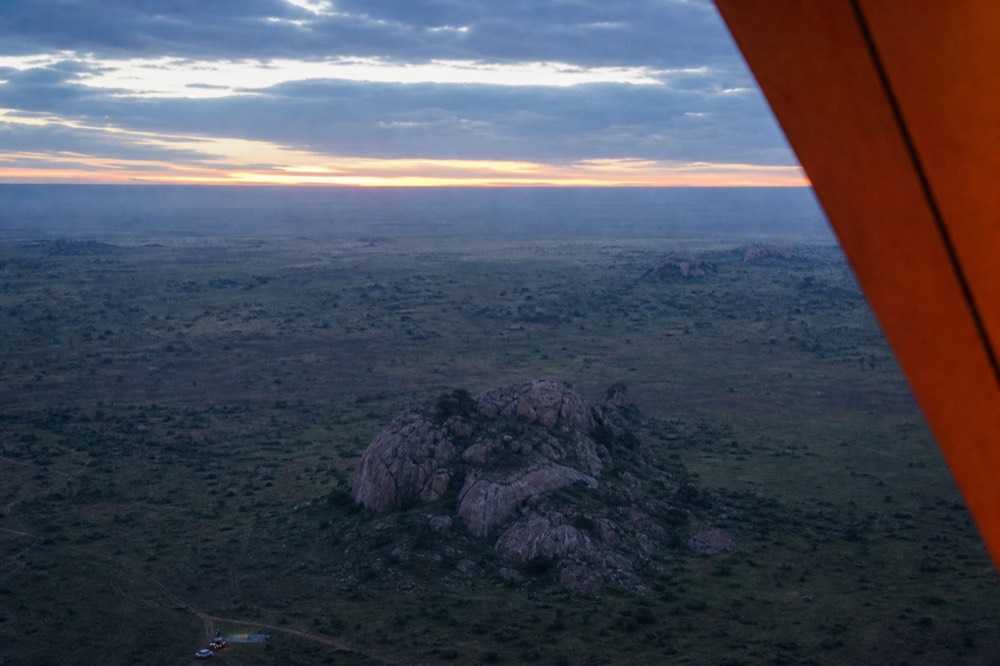 Flat open plains seen from altitude at dawn, long shadows thrown by scattered granite outcrops, the curved edge of a warm orange balloon envelope in frame