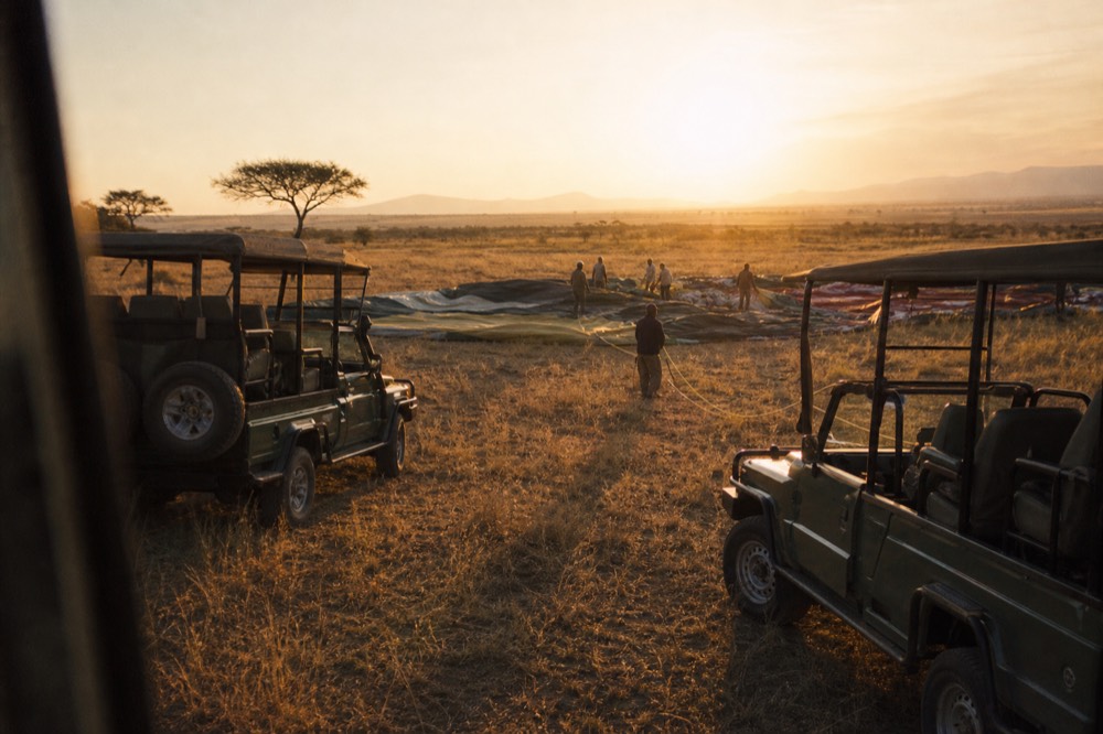 Two safari vehicles waiting on a grass clearing beside deflated balloon fabric spread flat, ground crew coiling ropes, low golden sun behind