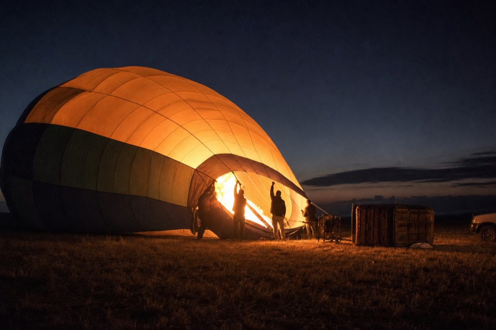 Balloon envelope half inflated before sunrise, fabric glowing warm from the burner inside, ground crew silhouetted holding the mouth open against a dark sky