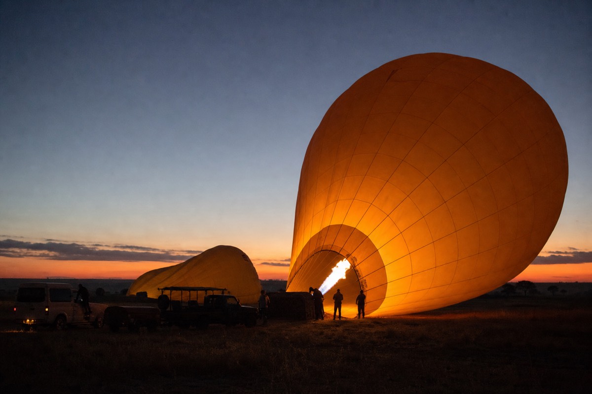 A balloon crew firing the burners during pre-dawn inflation on the savanna, envelope glowing against a dark orange horizon
