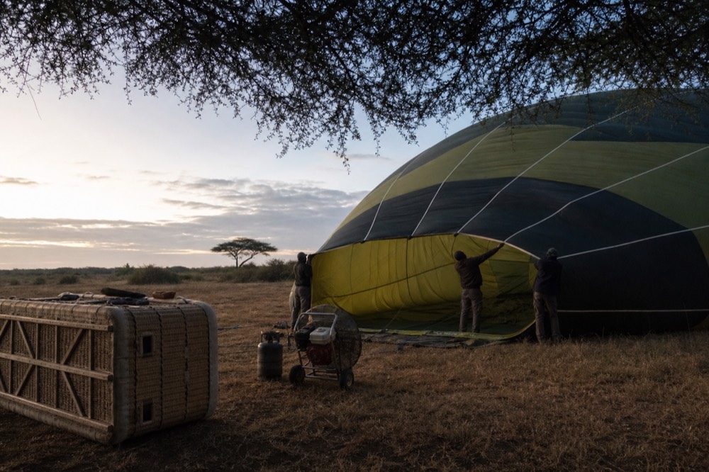 Balloon envelope inflating on flattened grass before sunrise, ground crew holding the mouth open while a fan drives cold air inside