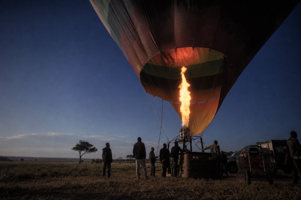 Propane burner throwing a tall orange flame into a balloon mouth against a deep blue pre-sunrise sky, crew silhouetted below the basket