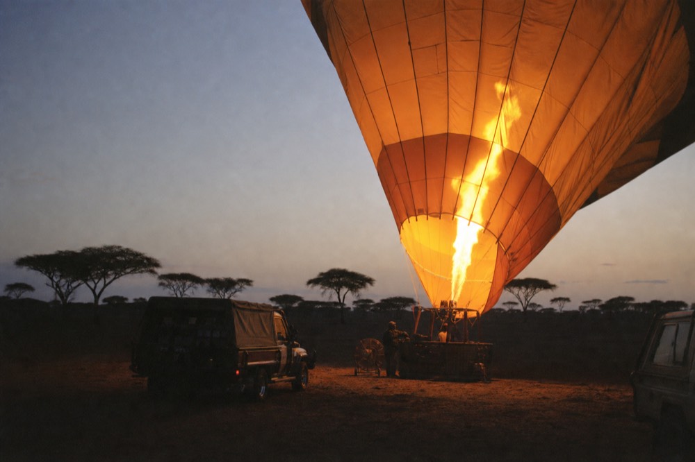 Burner flame throwing orange light up into the open throat of a warm-coloured balloon envelope on a dark launch field at dawn