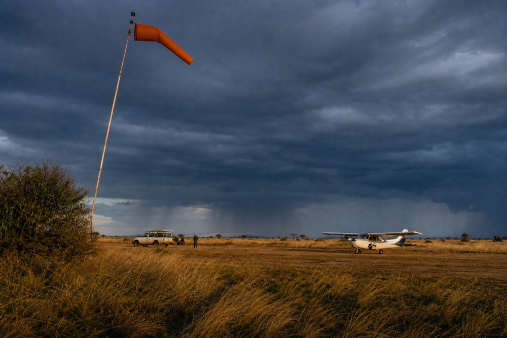 Wind-flattened yellow grass and a stiffened orange wind sock on a leaning pole beside a bush airstrip under a grey overcast dawn