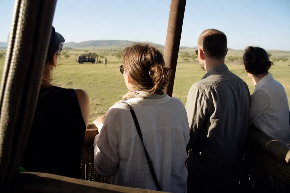 Four passengers standing shoulder to shoulder inside a padded wicker basket compartment, hands on braided rope handles, low sun across their shoulders