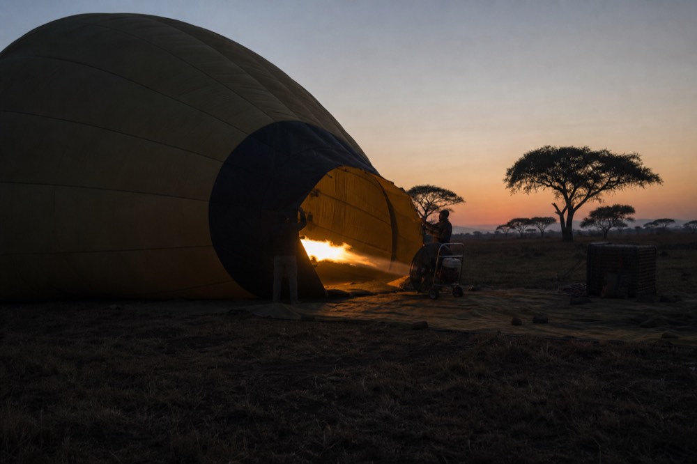 A balloon envelope half inflated on dark grass before sunrise, cold blue light, ground crew silhouettes holding the mouth open as the fan runs