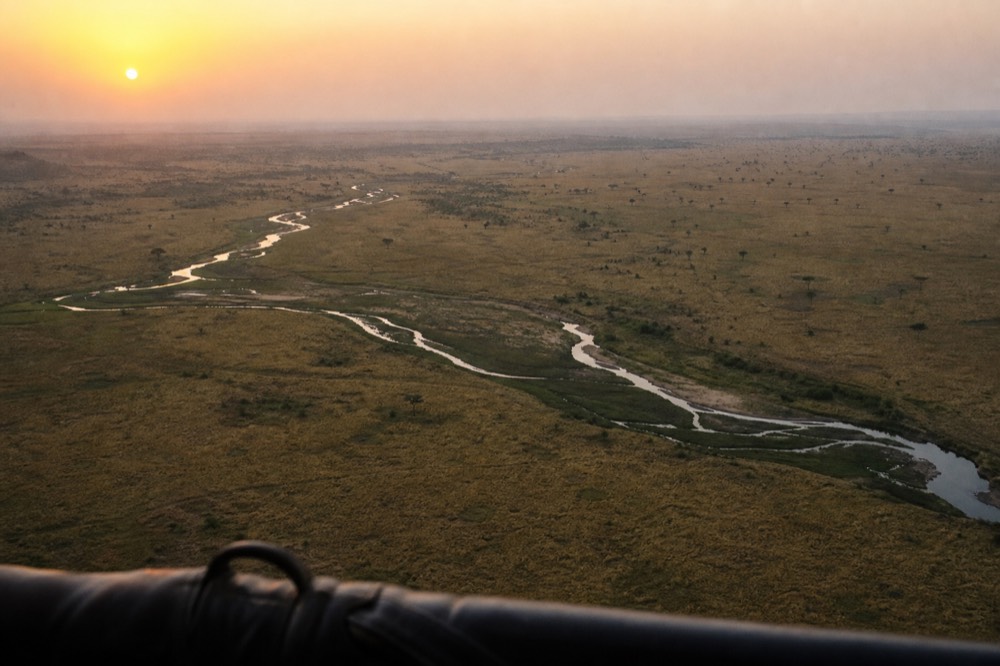 Aerial view of unbroken golden plains stretching to a hazy horizon at sunrise, a braided river cutting a dark line across the grass, no roads visible