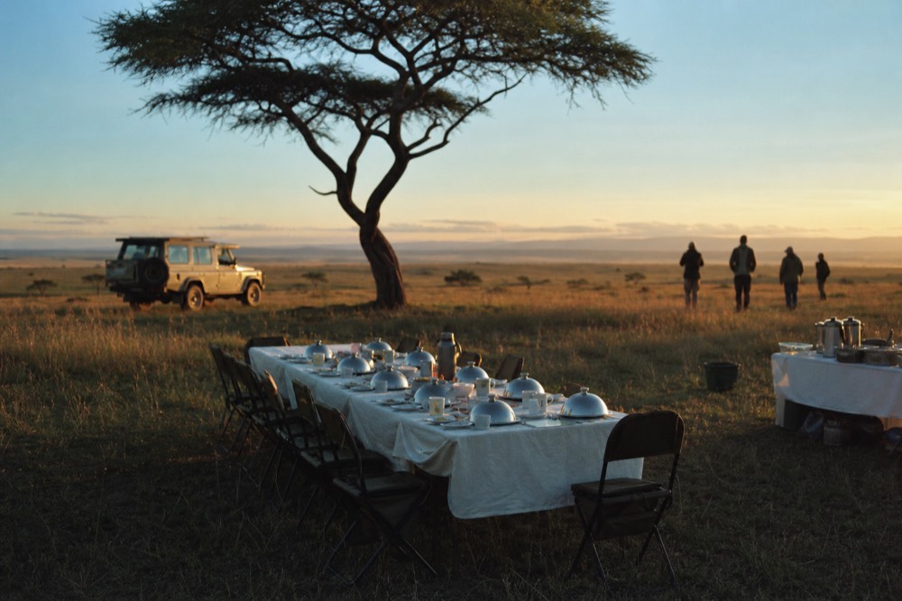A long breakfast table laid with white linen, silver domes and folding chairs standing in open grass under a flat-topped acacia in warm morning sun