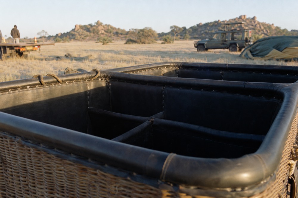 Empty padded compartments inside a wicker balloon basket at dawn, rope handles coiled on the rim, frost-grey grass and a pale horizon beyond