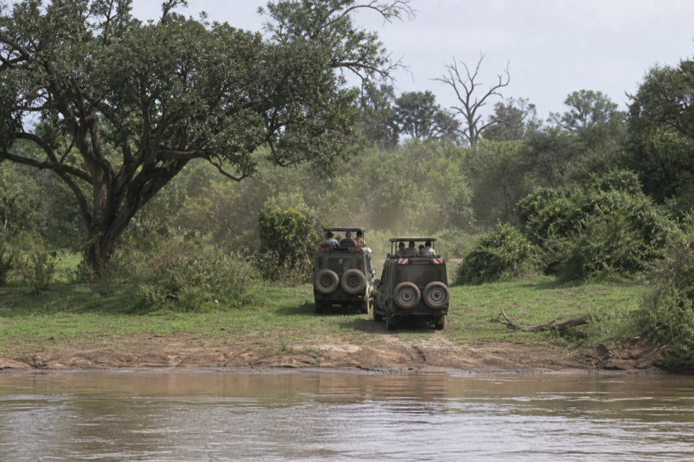 Two safari vehicles parked at a river bank at eye level with the brown water, heat shimmer and dust hanging above the roofs