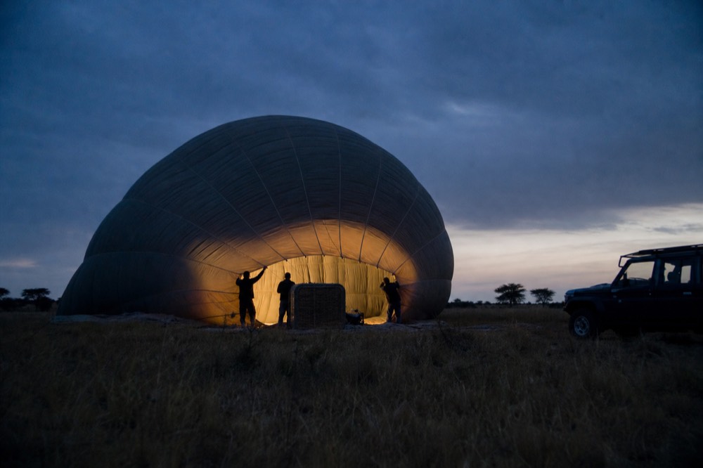 Pre-dawn balloon envelope half inflated on flattened grass, ground crew silhouettes holding the mouth open, cold blue light before sunrise
