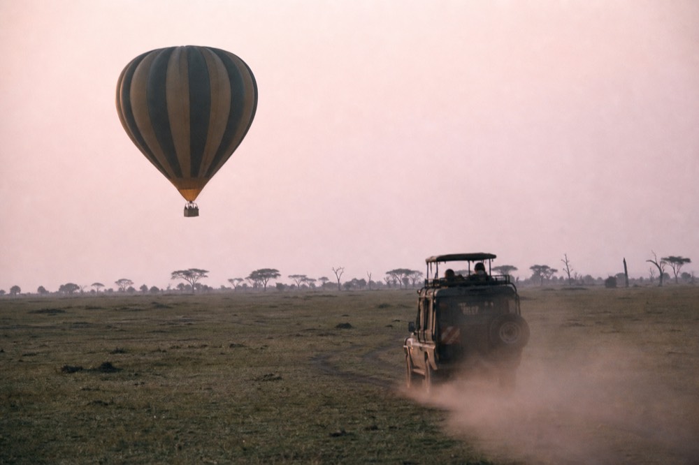 Chase vehicle following a low-flying balloon across open grassland, a pale plume of dust trailing behind it in early morning light