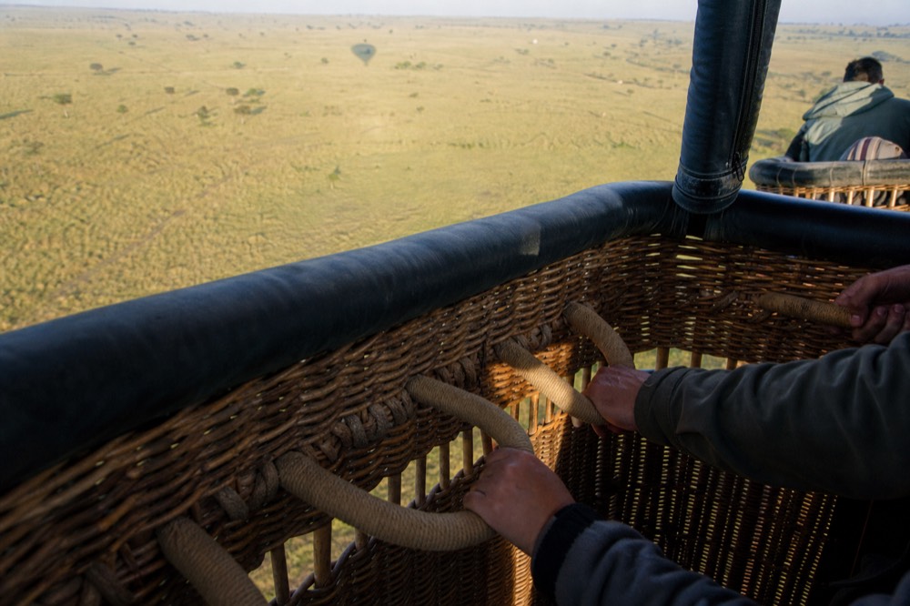 Hands gripping padded rope handles set into the inside of a wicker basket wall, pale grassland far below beyond the rim
