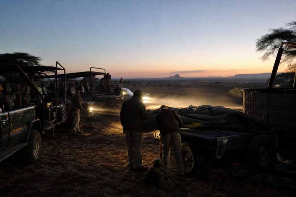 A line of open safari vehicles parked in darkness beside a flat launch field, headlights raking through dust, crew unloading a folded envelope from a trailer