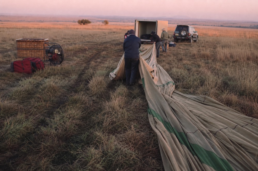 Landing drag marks torn through tall pale grass, a deflated envelope stretched out flat and crew rolling it toward an open trailer