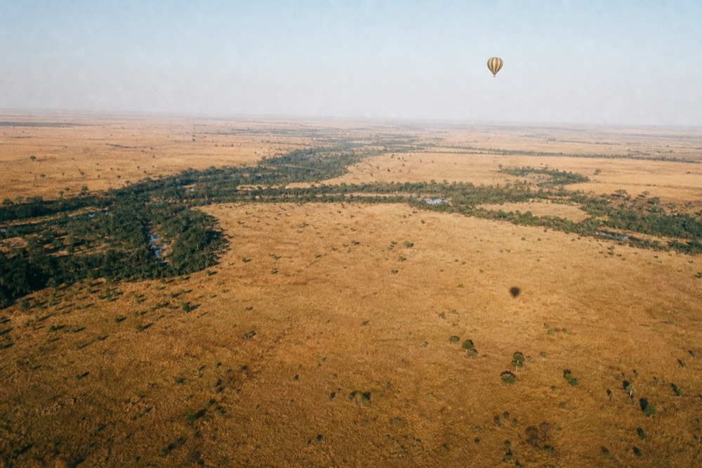 A single balloon high above a mosaic of burnt-orange grassland and dark ribbons of riverine trees, its shadow a small dot on the plain far below
