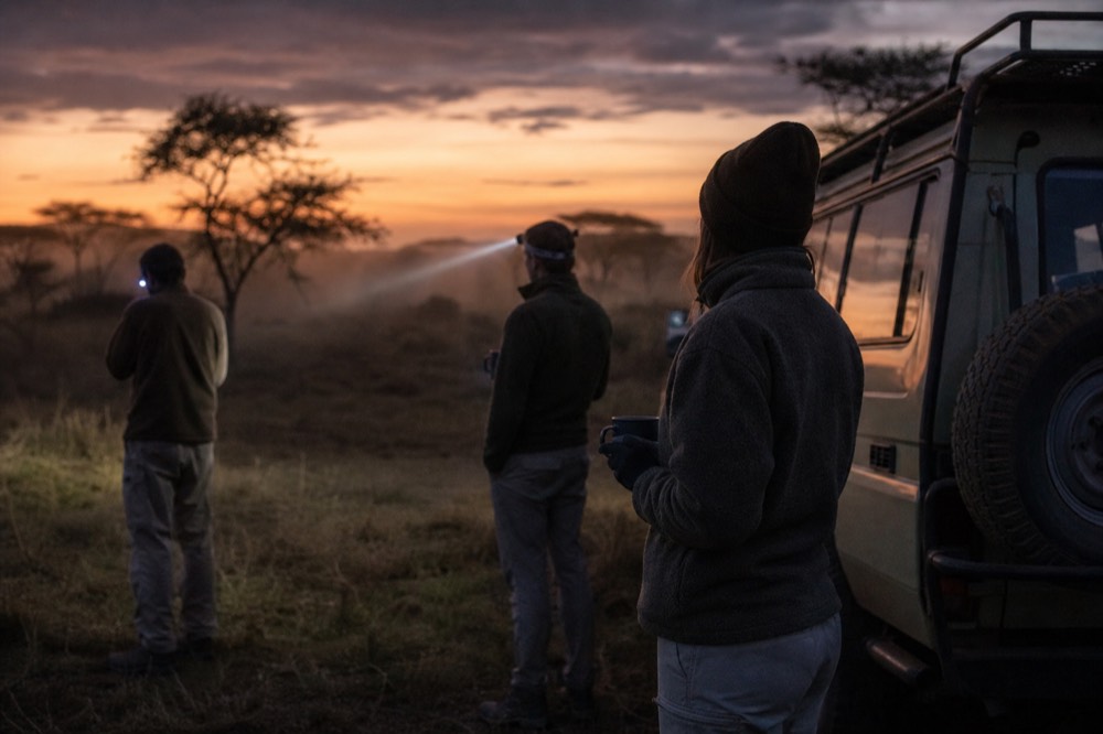 Passengers in fleeces standing beside a parked safari vehicle on a dark grass field, gripping enamel mugs, headlamp beams crossing in the cold air