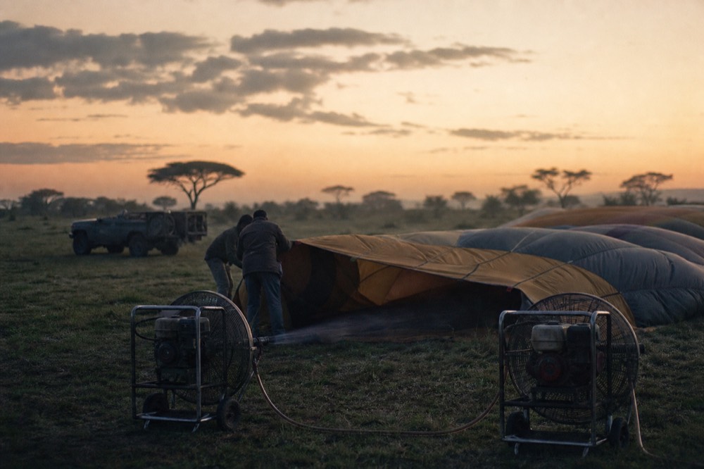 Two industrial petrol fans blowing cold air into a limp balloon envelope stretched flat across dewy grass, crew members holding the fabric mouth open