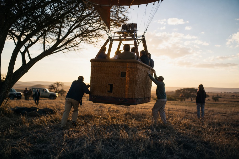 The moment of lift-off, a wicker balloon basket a hand's width above flattened grass with crew hands releasing the rim, low orange sun behind