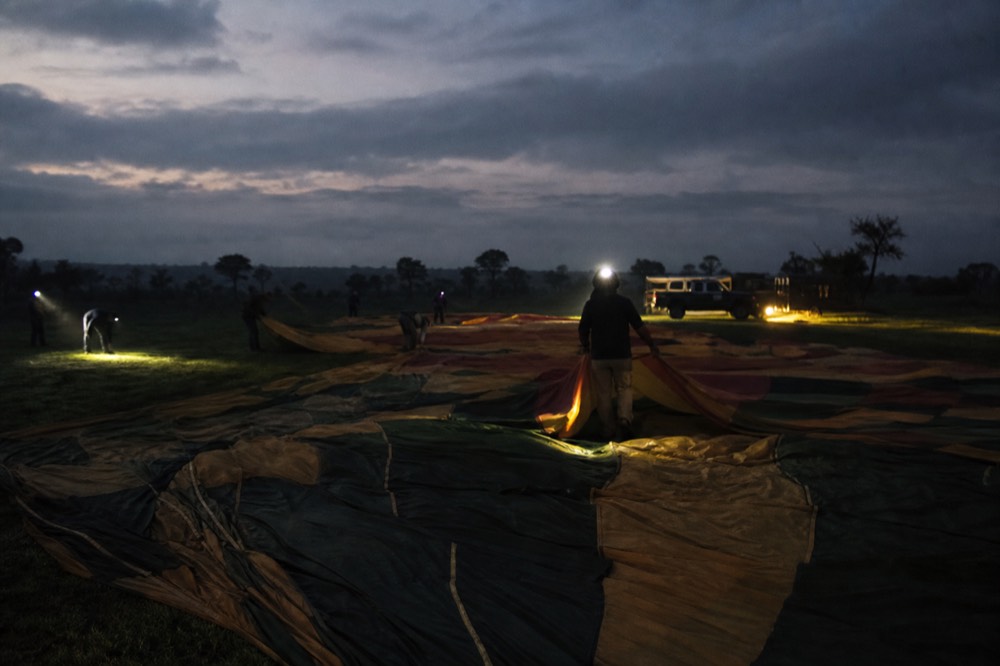 Balloon fabric spread flat on dark dew-covered grass before sunrise under headlamp beams and cold blue light, ground crew silhouettes stretching the panels out