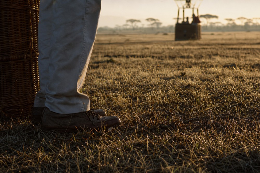 Worn brown closed walking shoes standing ankle-deep in dew-soaked pale grass beside the woven base of a wicker basket, early sunlight raking across the ground