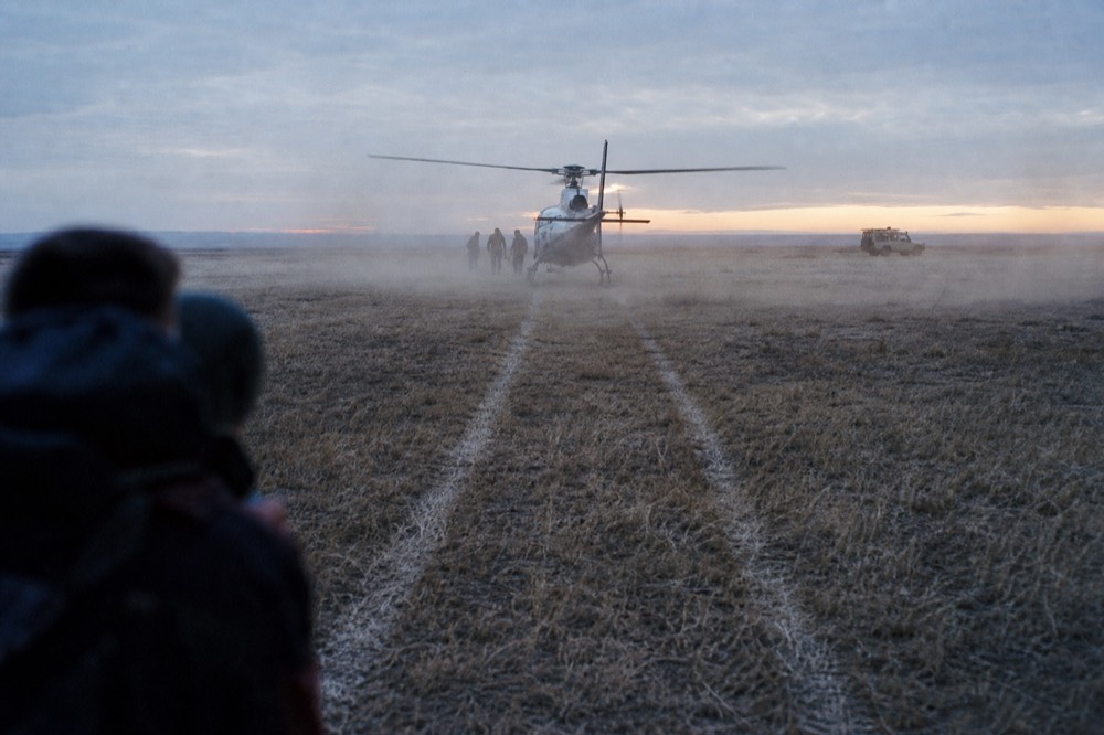 Two parallel skid marks pressed into pale dry grass at the landing spot with dust still hanging in low sunlight and a vehicle waiting in the distance