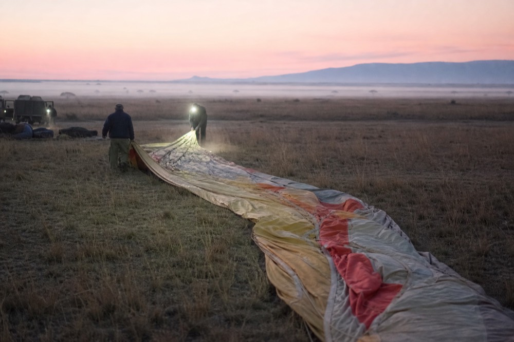 Ground crew unrolling a folded balloon envelope across flattened grass in cold pre-dawn light, headlamp beams cutting through low dust