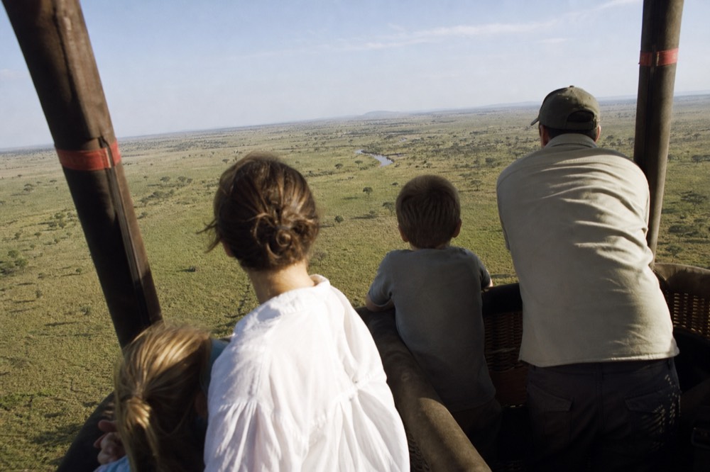 A family group standing in a divided balloon basket photographed from behind at low altitude, heads turned toward grassland far below