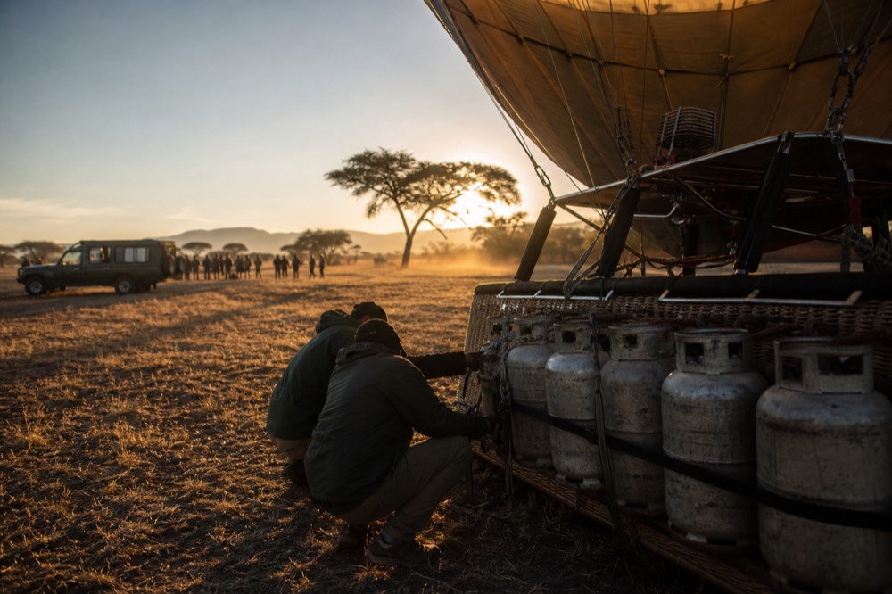 Ground crew crouched beside a basket checking strapped propane cylinders, low dawn light raking across flattened grass